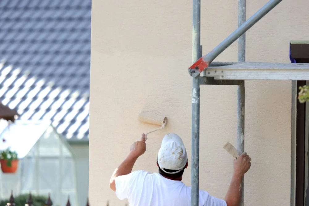 Maler im weißen T-Shirt rollt hellen Außenputz mit Farbrolle auf Hauswand – Fassadenanstrich