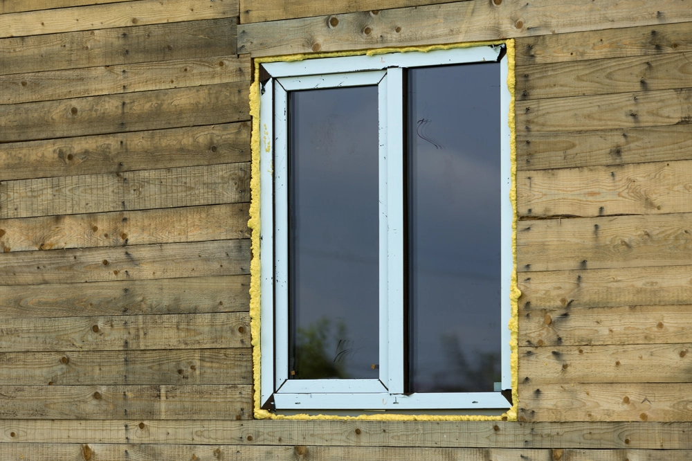 Fenstermontage in Holzfassade mit sichtbarer gelber Montageschaumdämmung um weißes Fenster