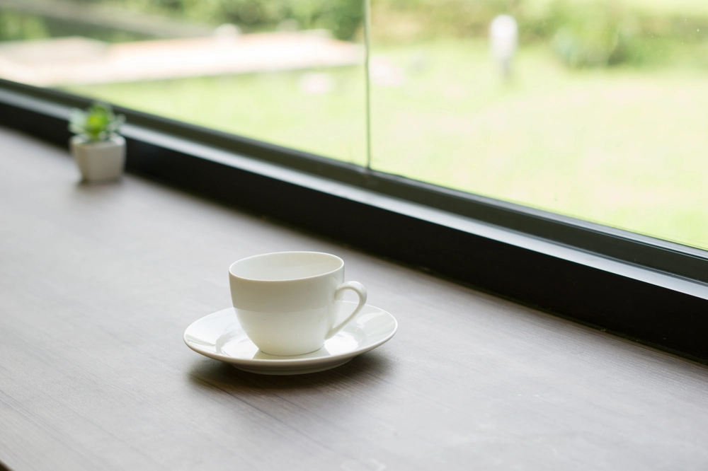 Weißer Kaffeebecher auf Holztisch vor einem dunklen Aluminiumfenster