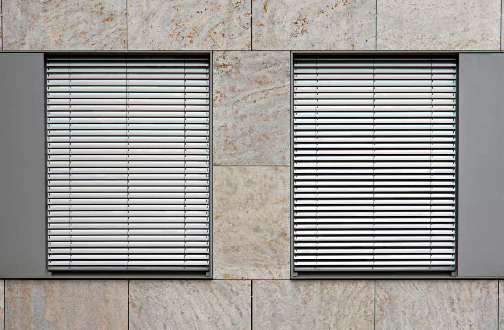 Zwei Fenster mit geschlossenen Außenjalousien in gleichmäßiger Natursteinfassade