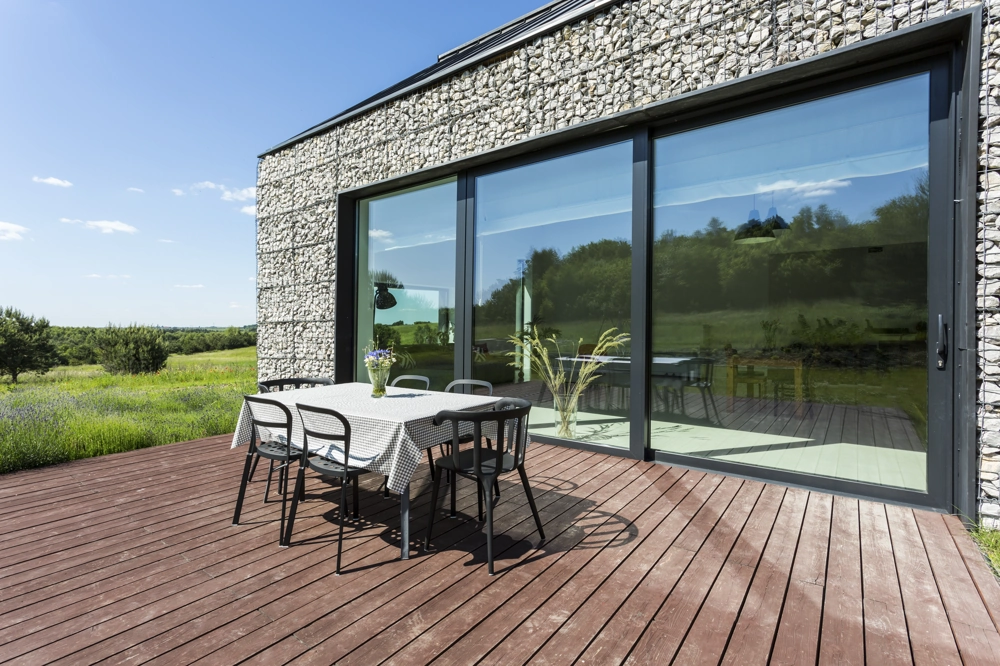 Holzterrasse mit Gartentisch und Stühlen vor Steinfassade und großen Aluminiumfenstern